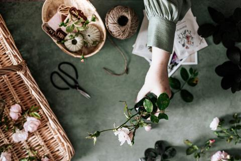 A woman's hand holding flowers out, surrounded by more cut flowers, a pair of shears, and a basket of twine.