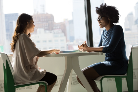 two women having a discussion at a round table placed by a larger window